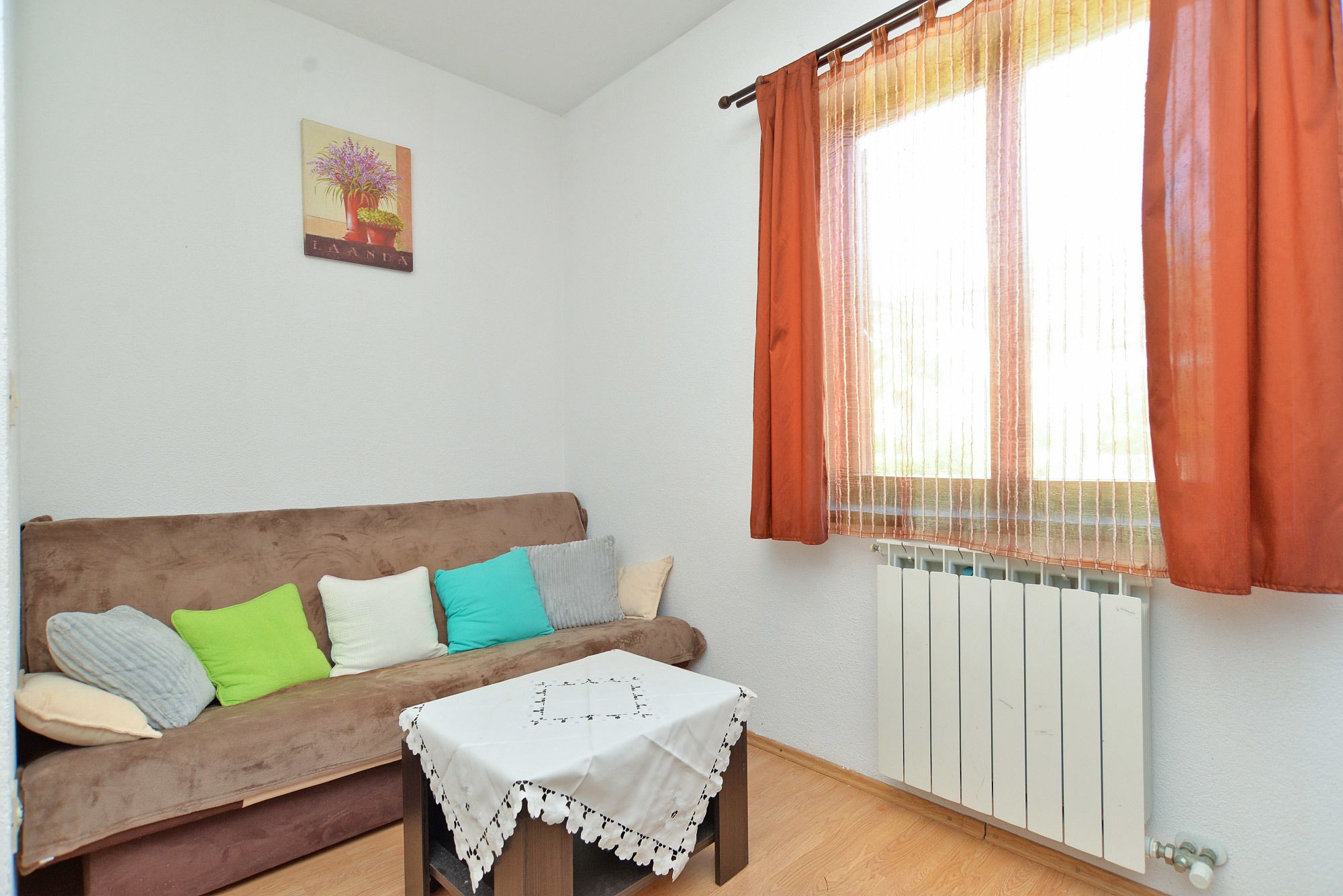 Un salon avec un canapé marron, une petite table recouverte d’une nappe blanche et une fenêtre ornée de rideaux orange.