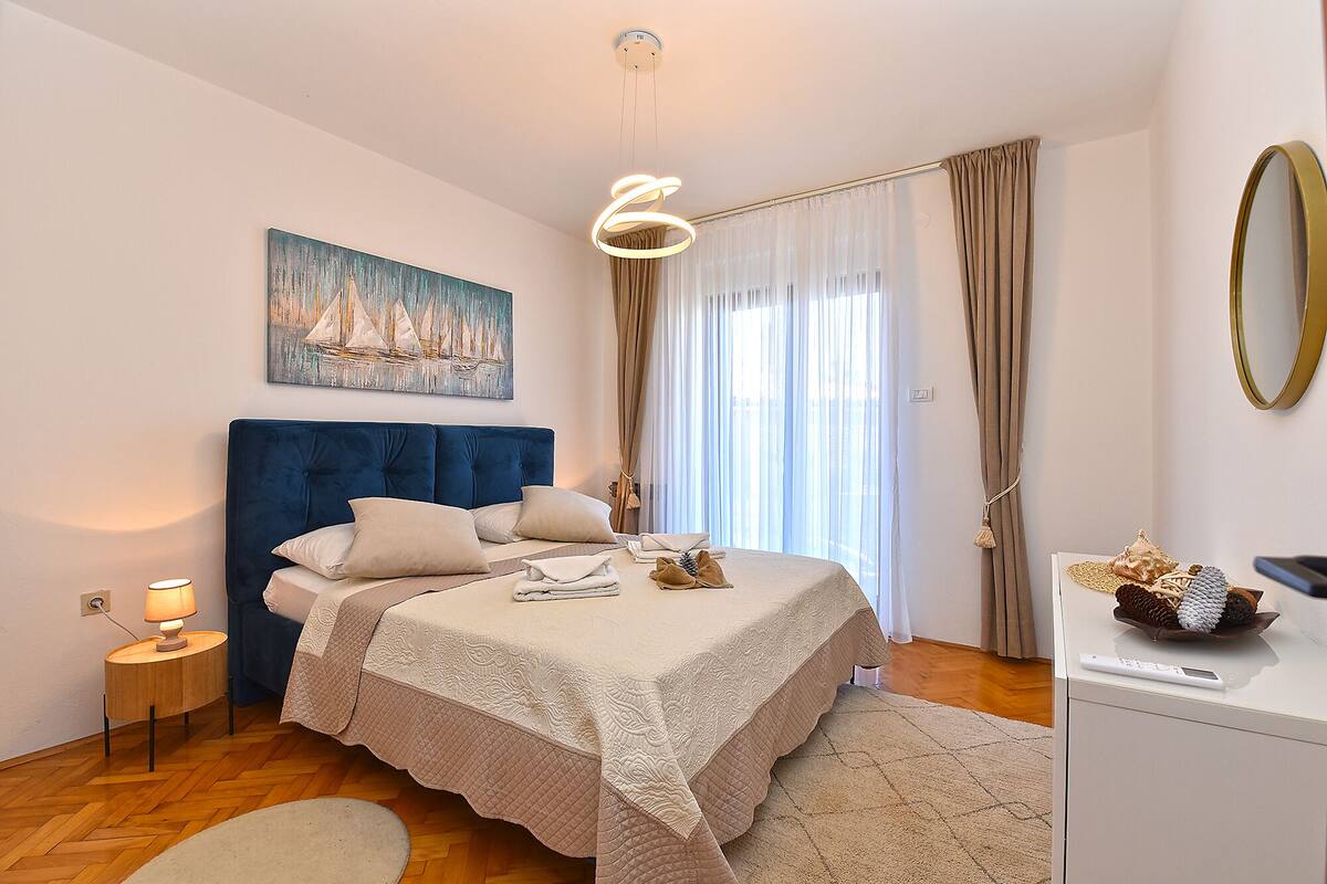 A bedroom with a large bed, a bedside table with a lamp, a painting on the wall, and a window with curtains.