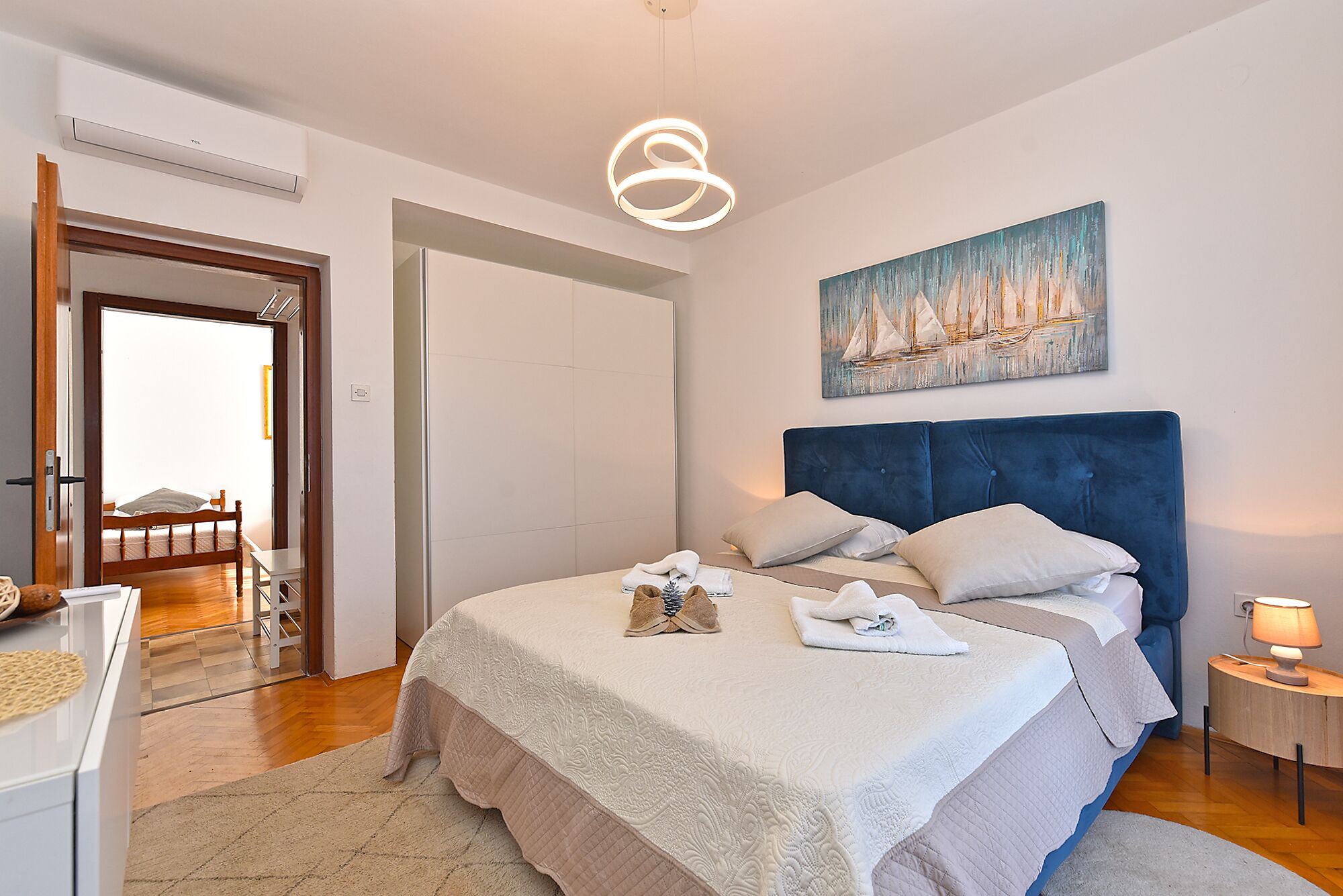 A bedroom with a bed, a painting, a bedside lamp, and a door leading to another room.