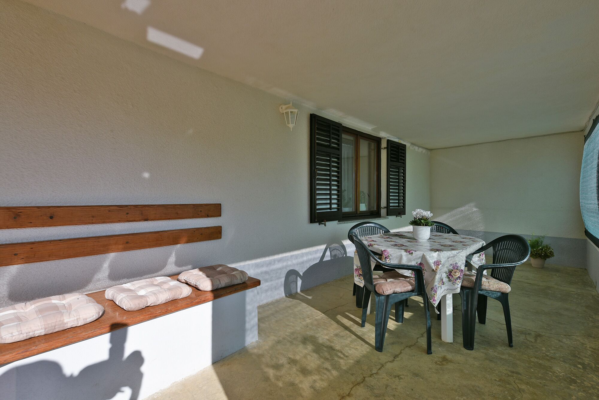 Un patio soleggiato con una panca di legno, un tavolo con sedie e una finestra con persiane.