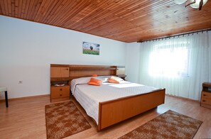 A bedroom with a wooden headboard, a bed with orange pillows, a nightstand, a bedside table, and a window with sheer curtains. - Apartment 1492 (Barban)