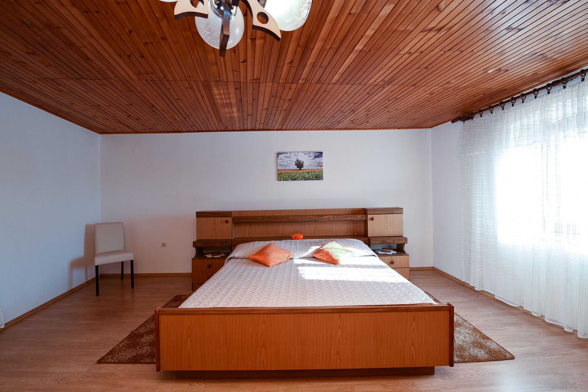 A bedroom with a wooden ceiling, a bed with orange pillows, a nightstand, and a chair.