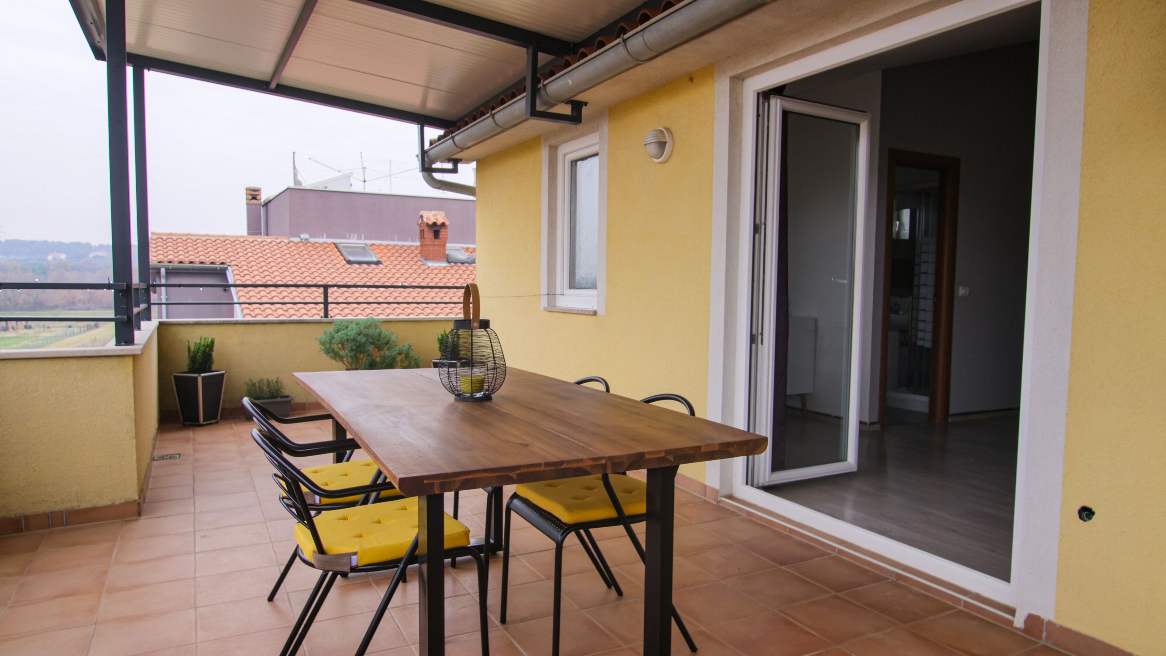 Une terrasse avec une table en bois et des chaises jaunes, une petite plante en pot et une vue sur le paysage urbain.
