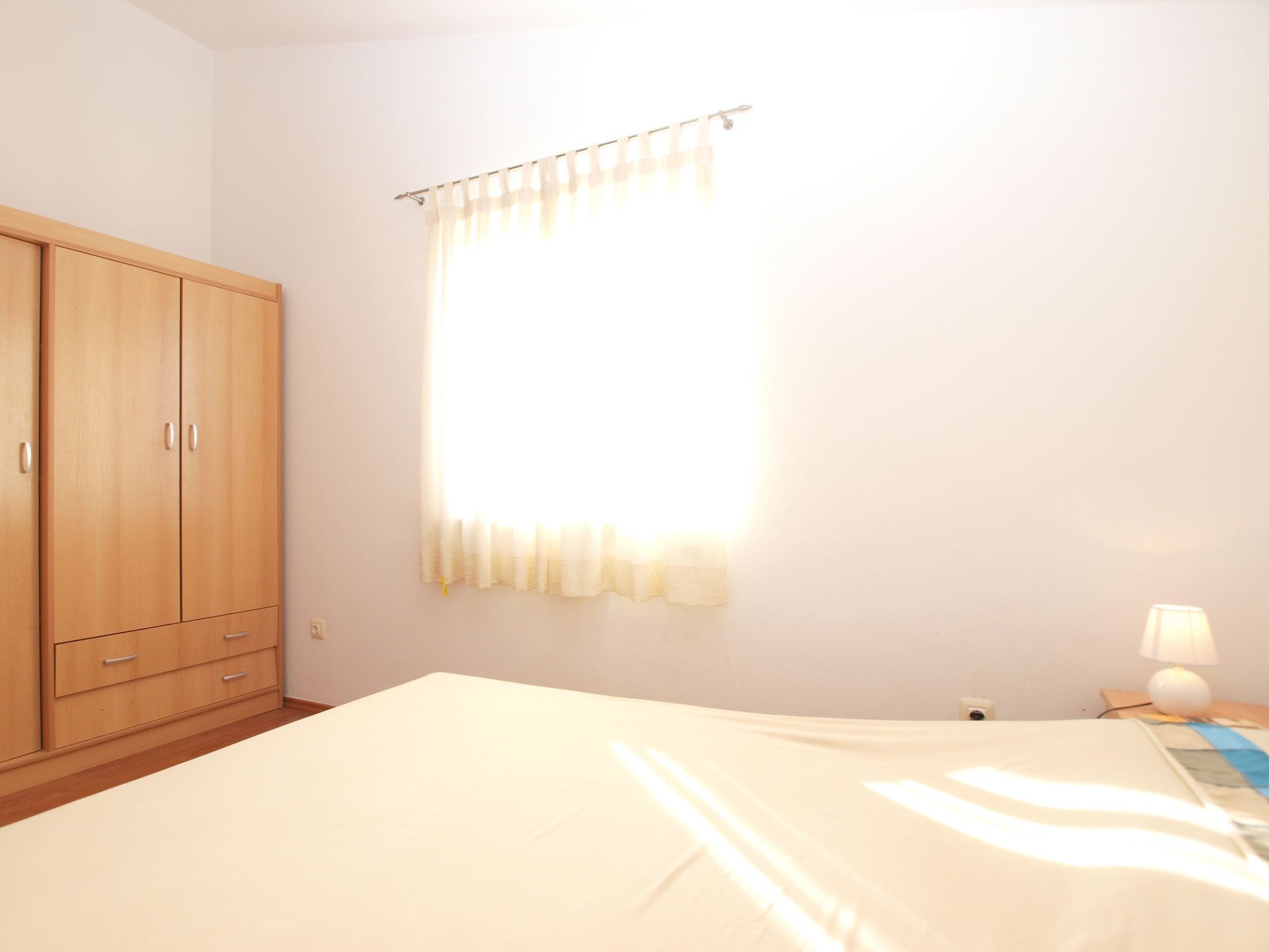 A bedroom with a bed, a wooden wardrobe, a window with curtains, and a bedside lamp.