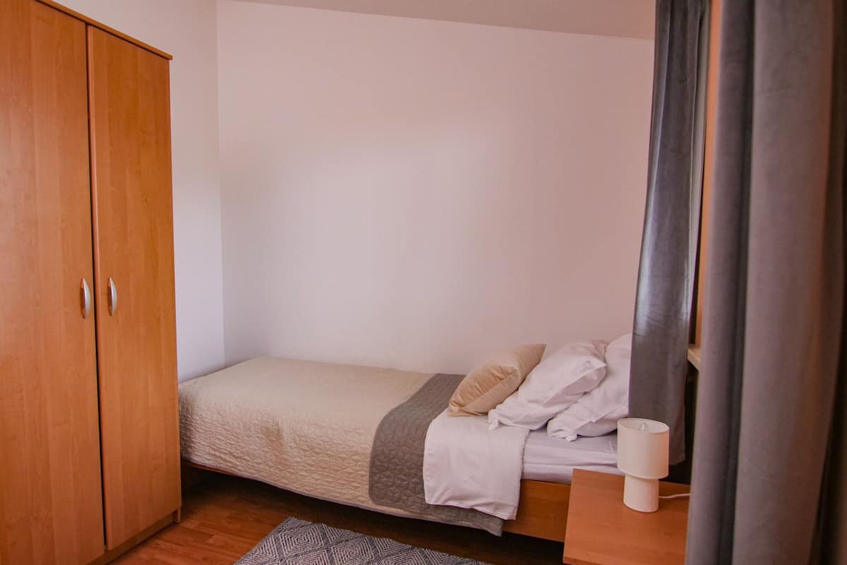 Une petite chambre avec un lit simple, une armoire en bois, une table de chevet avec une lampe et un tapis gris.
