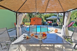 A covered outdoor area with a pool, lounge chairs, and a table set with food and drinks. - Villa 1505 (Liznjan)