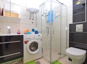 A compact bathroom with a washing machine, a glass shower enclosure, and a toilet. - Apartment 1507 (Fazana)