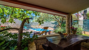 An outdoor patio with a stone wall, a pool, lounge chairs, and a wooden table with a fruit basket. - Apartment 1510 (Marcana)