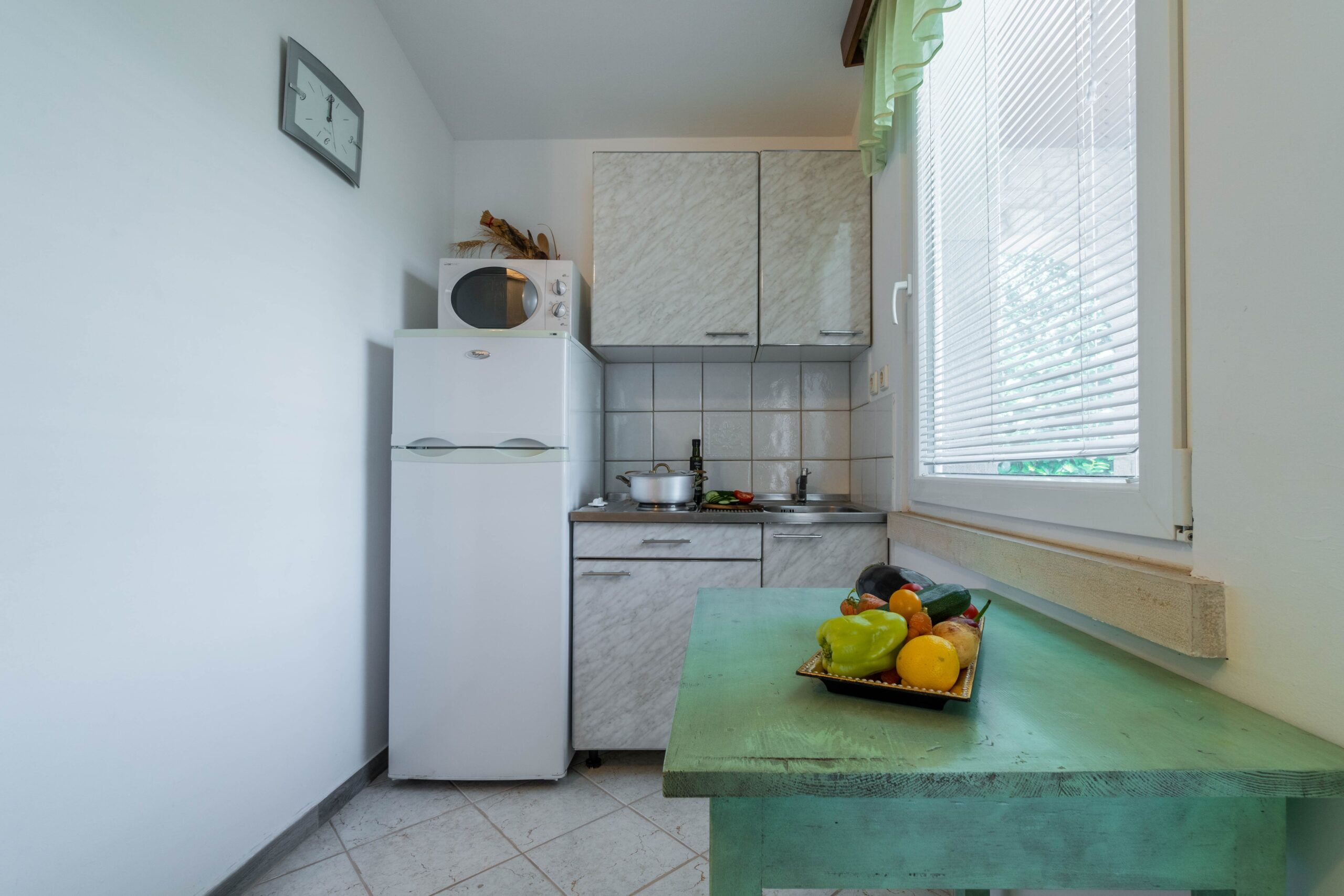 A compact kitchen with a refrigerator, microwave, and tiled backsplash. A window with blinds is visible, and a table with a fruit basket is in the foreground.