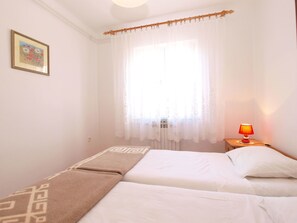 Ein Schlafzimmer mit zwei Betten, einem Fenster mit durchsichtigen Vorhängen, einer Nachttischlampe und einem gerahmten Bild an der Wand.