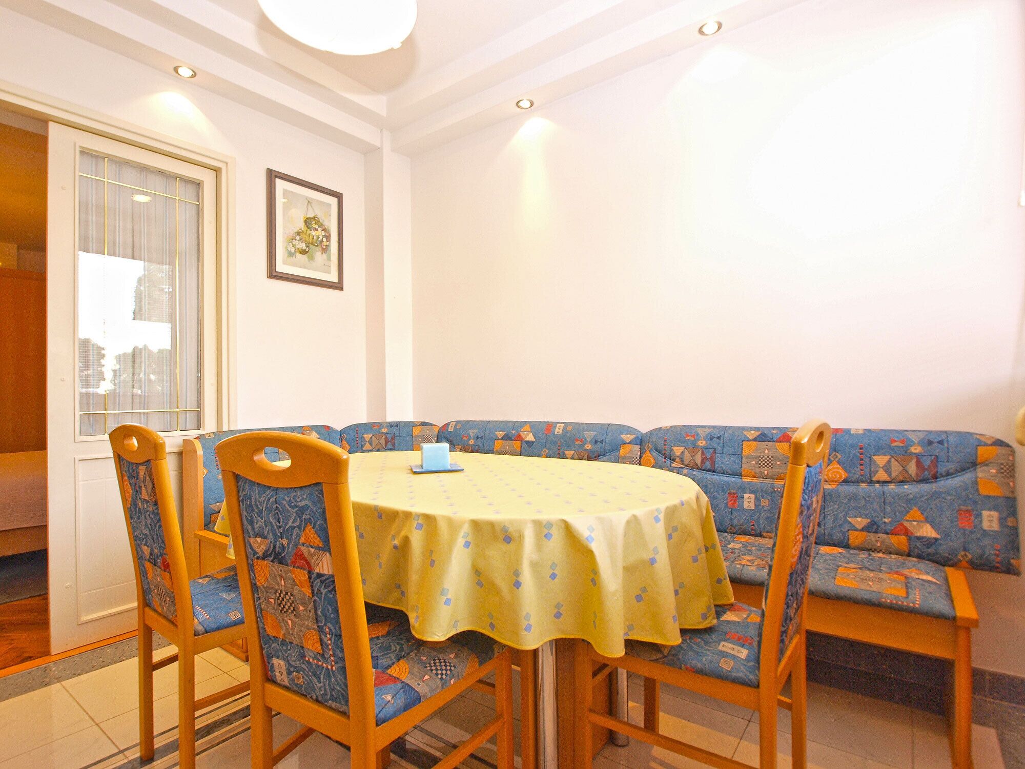A dining area with a round table covered by a yellow tablecloth, surrounded by four chairs with patterned cushions.