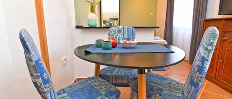 A dining area with a round table, two patterned chairs, a vase with flowers, a bowl of fruit, and a mirror above the table.