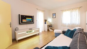 Een moderne woonkamer met een flatscreen televisie aan de muur, een wit bureau met een stoel, een bank met kussens en een raam met gordijnen.