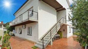 Um edifício de dois andares com uma varanda no andar superior, uma escadaria que leva ao nível superior e uma pequena área externa com mesa e cadeiras.