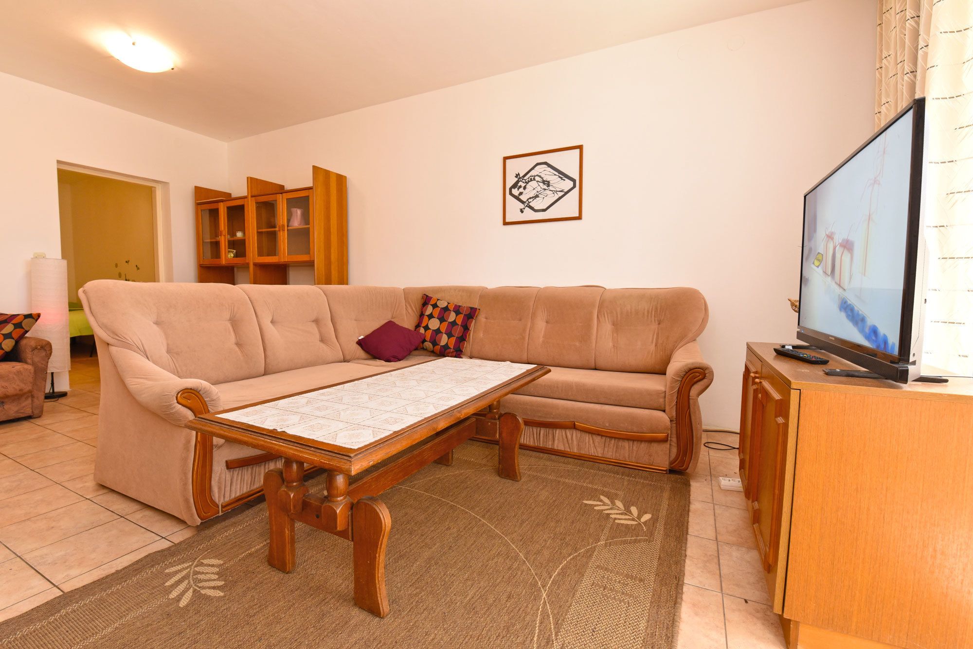 Een woonkamer met een beige bank, een houten salontafel, een televisie op een houten dressoir en een vloerkleed met een patroon.