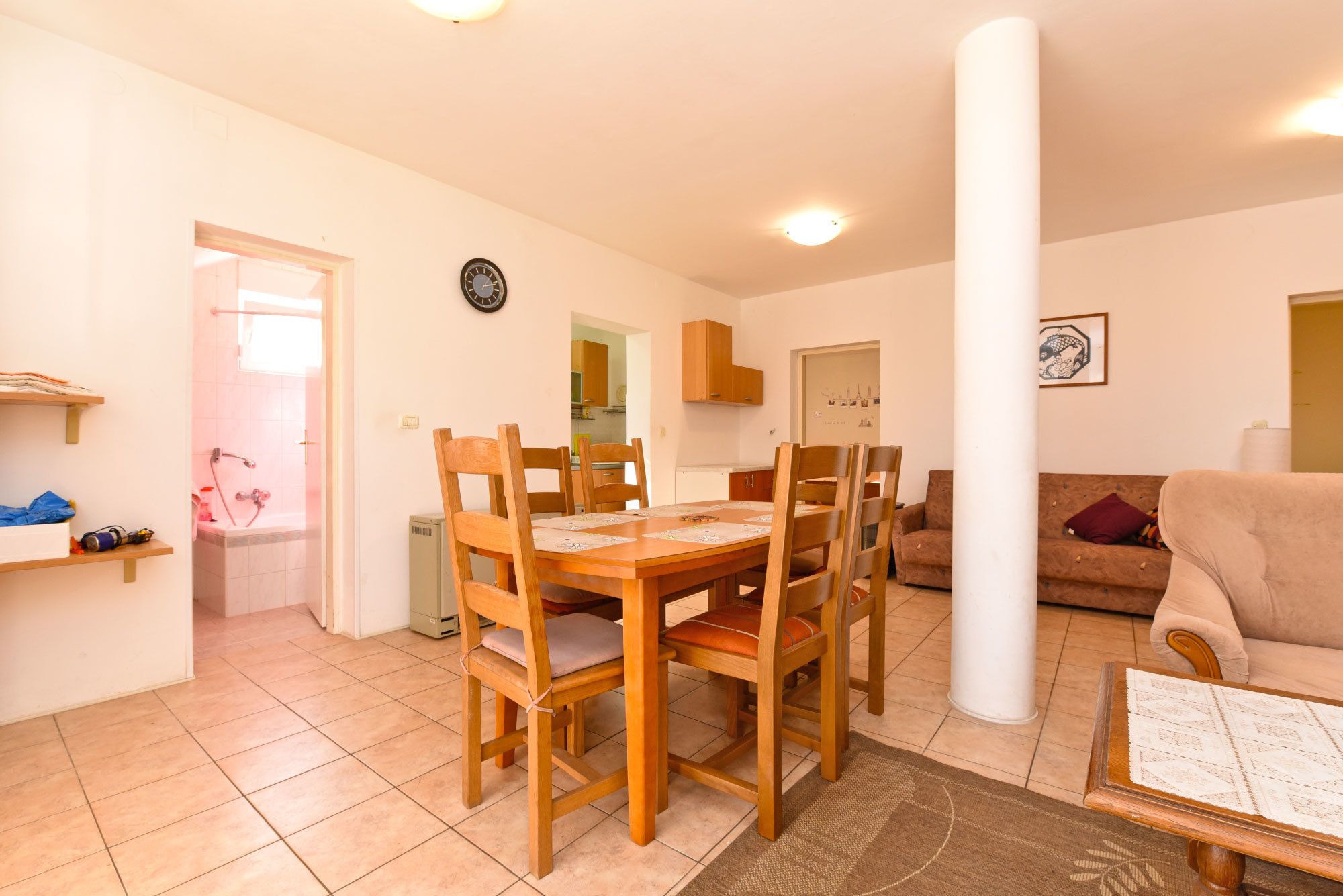 Ein Essbereich mit Holztisch und -stühlen, eine Küche im Hintergrund und ein Wohnzimmer mit Sofa und Lampe.