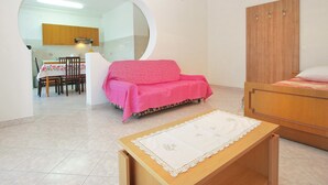 A room with a pink sofa, a wooden bed, a small table with a lace tablecloth, and a kitchen in the background. - Apartment 1563 (Pula)