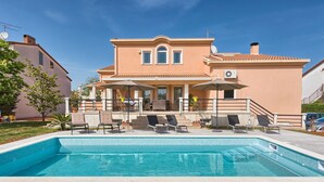 Una casa de dos pisos con piscina, mobiliario exterior y un patio cubierto.
