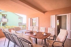 A covered patio with a wooden table set for four, surrounded by wicker chairs with cushions.