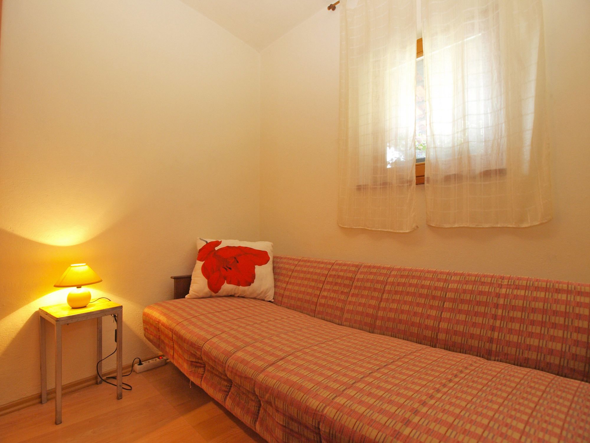 A small, single-bed room with a plain wall, a bedside table with a lamp, a red pillow, and a window with sheer curtains.