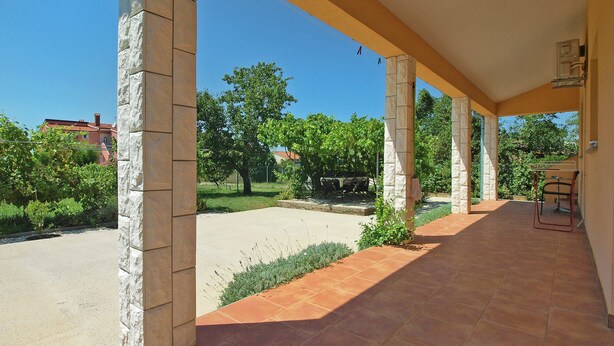 A covered outdoor area with stone pillars, a tiled floor, and a view of greenery and buildings in the background. - Apartment 1573 (Fazana)