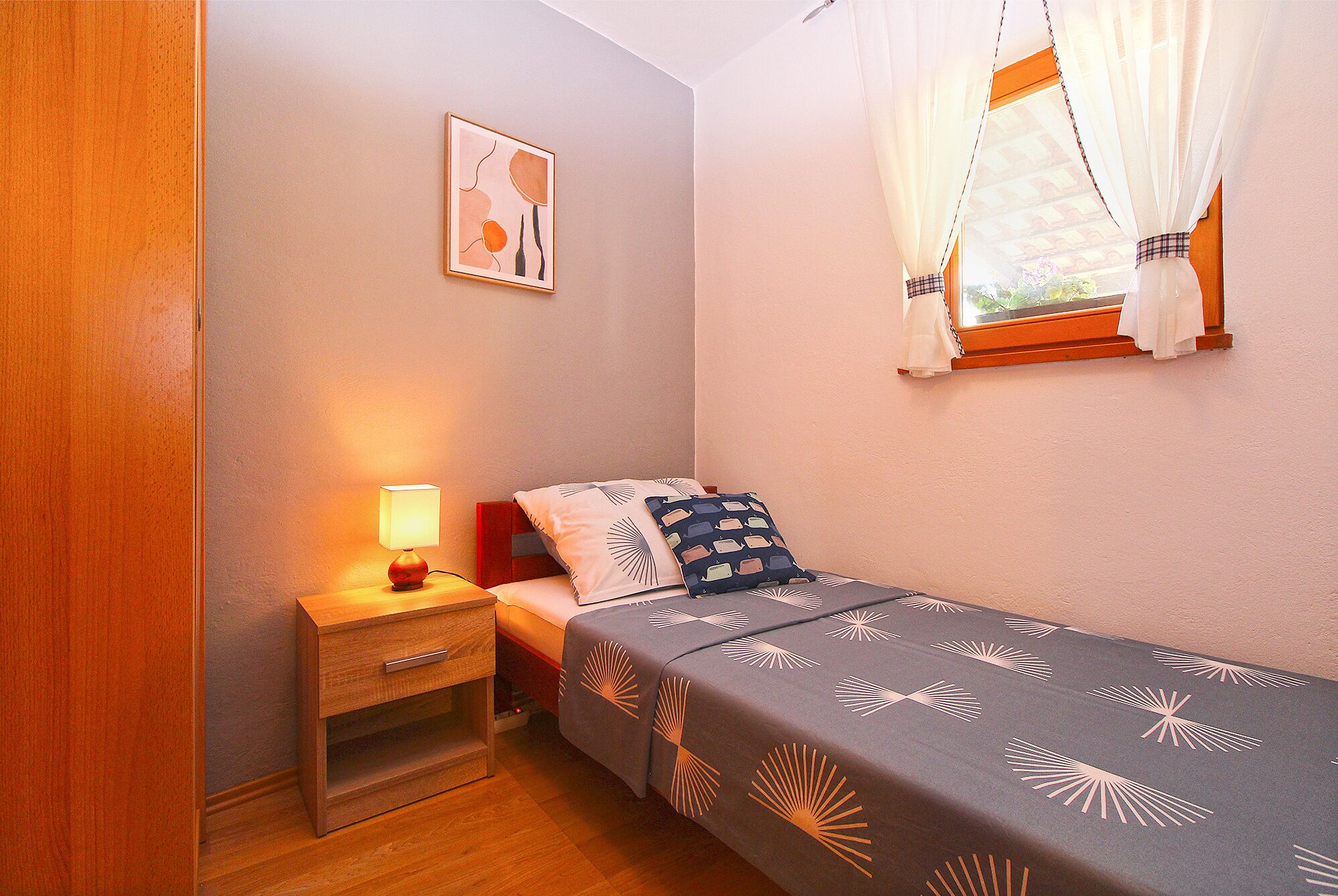 A small bedroom with a single bed, a nightstand with a lamp, a framed picture on the wall, and a window with curtains.