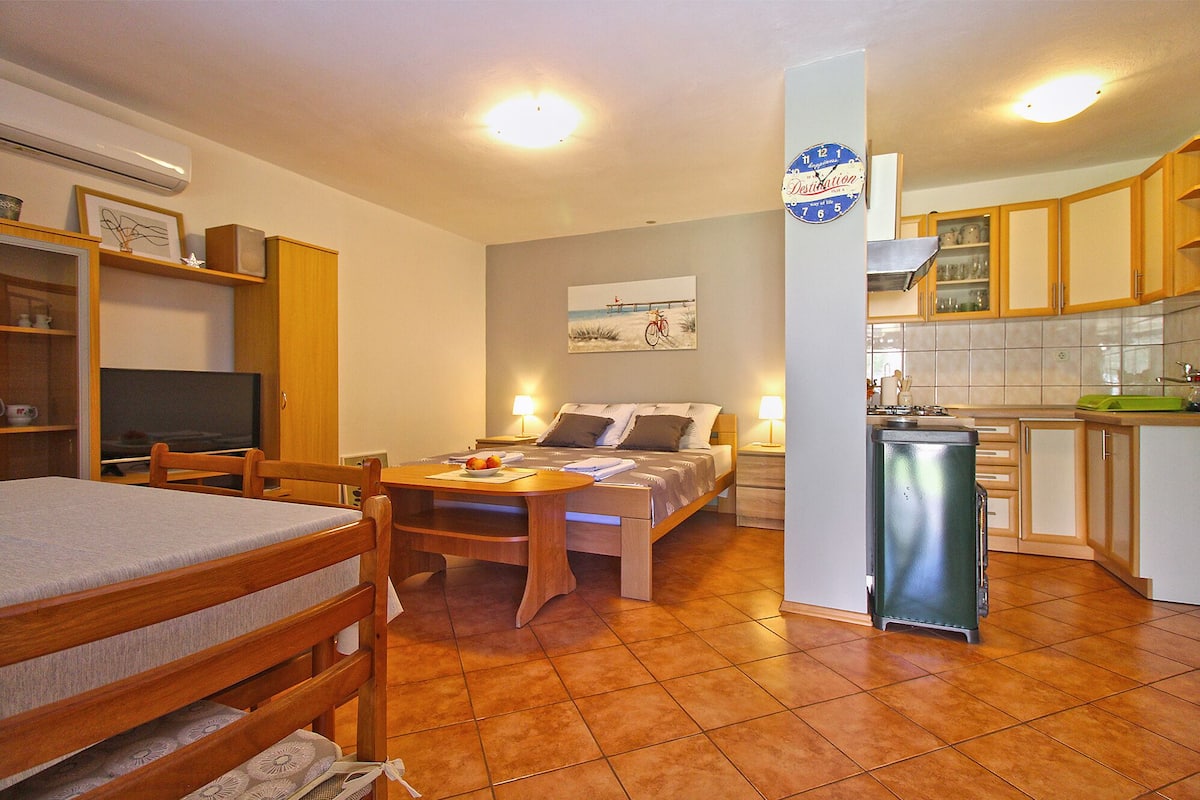 A compact room with a bed, a desk, a chair, a TV, a lamp, and a kitchenette.