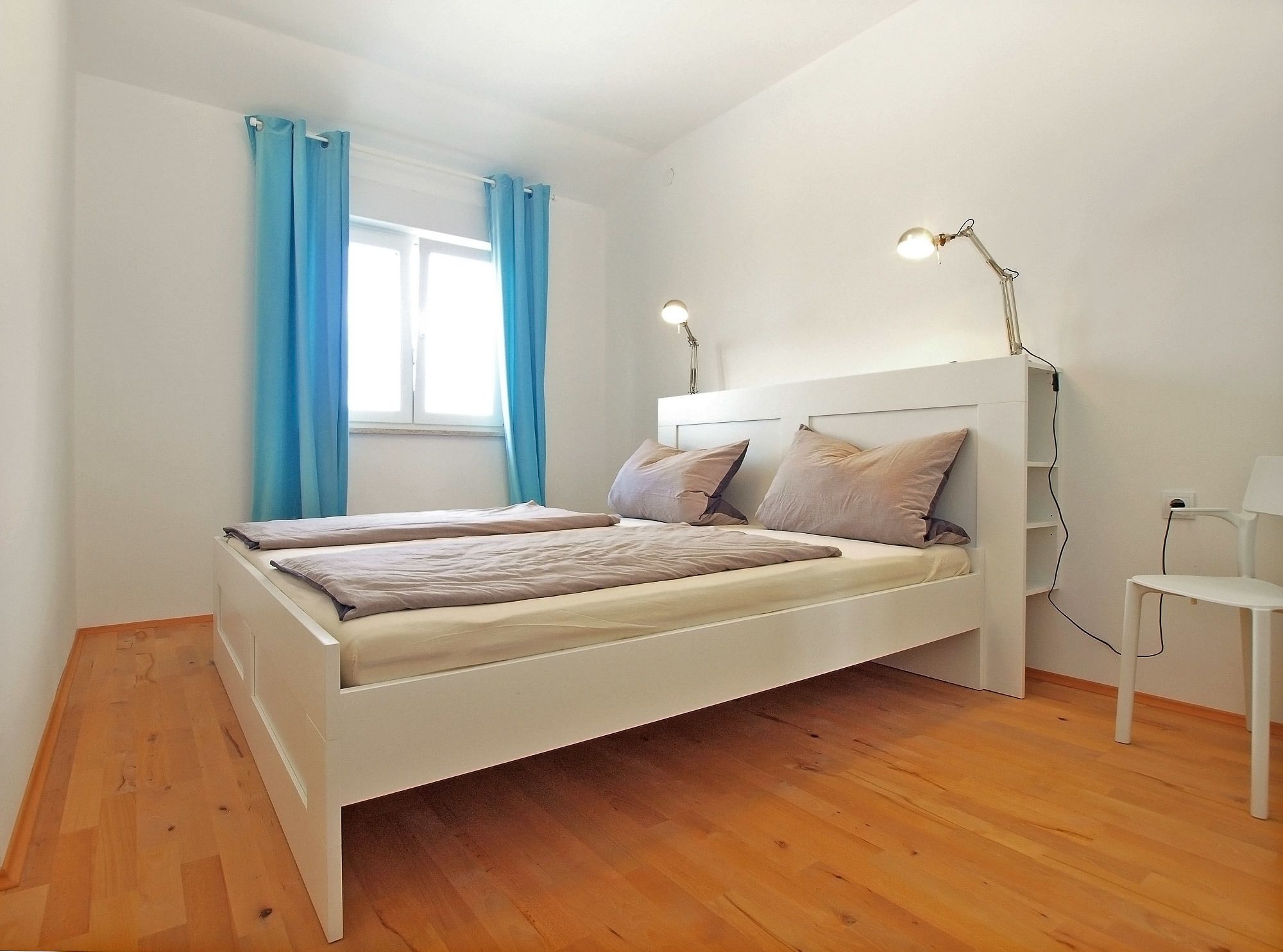 A bedroom with a white bed, two bedside lamps, and a window with blue curtains.