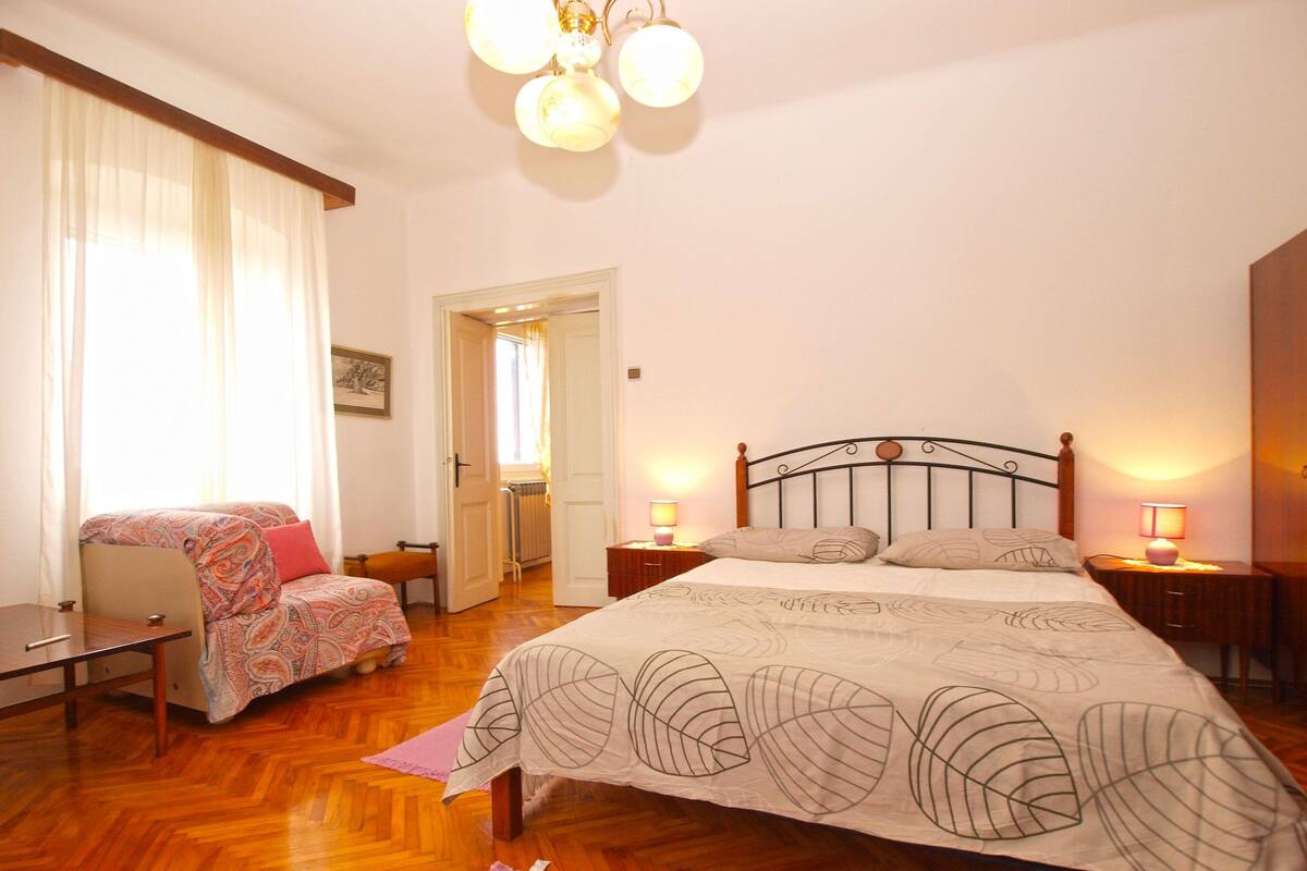 A bedroom with a bed, bedside tables, a chair, a window with curtains, and a door leading to another room.