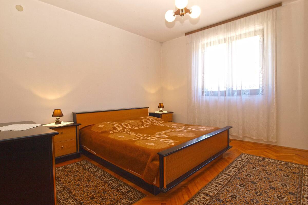 A bedroom with a wooden bed, two bedside tables with lamps, a large window with sheer curtains, and a patterned rug on the floor.