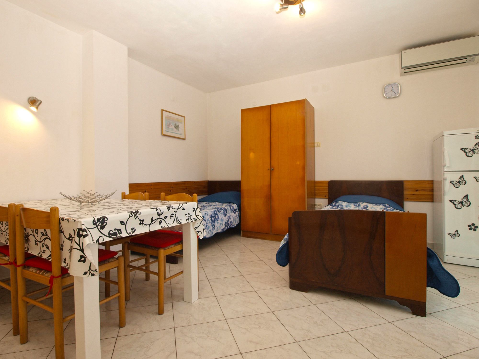 Een kleine kamer met een eettafel, twee eenpersoonsbedden, een kledingkast en een koelkast.