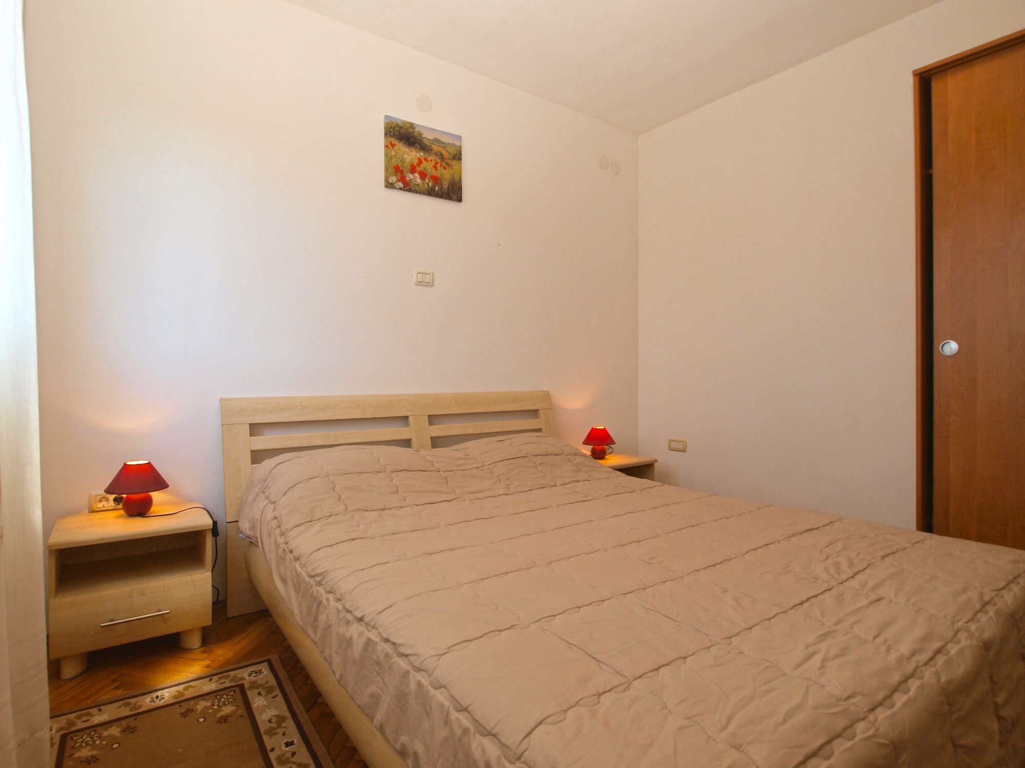 Une chambre avec un lit simple, une table de chevet avec une lampe, un petit tableau accroché au mur et une porte en bois.