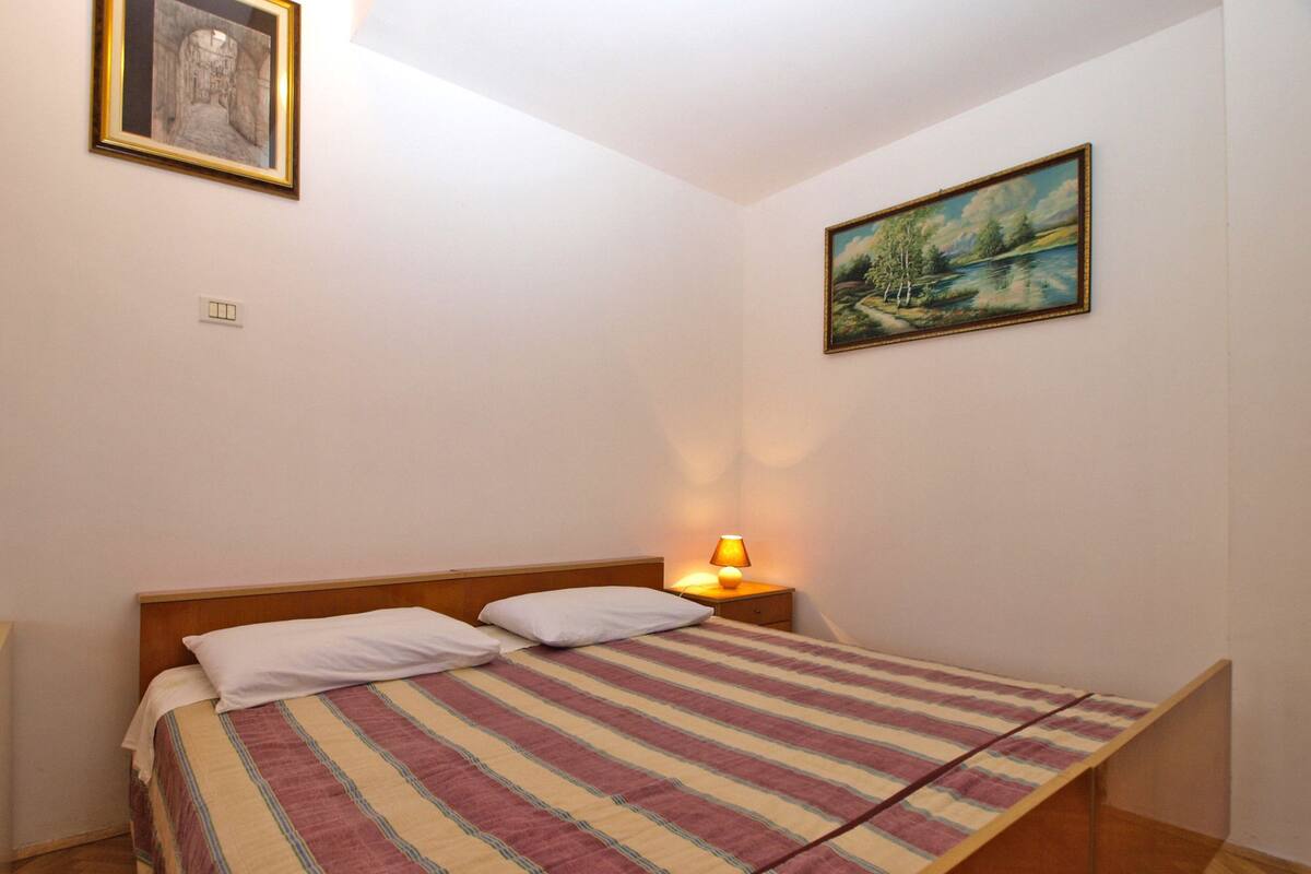 Un dormitorio con cama, mesita de noche con lámpara, un cuadro en la pared y una ventana.