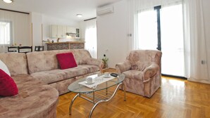 Een moderne woonkamer met een bank, fauteuil en glazen salontafel. Achtergrond is een keuken met witte kastjes en een betegelde achterwand.