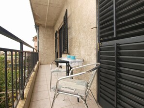 A balcony with a table and two chairs, a closed black shutter, and a view of greenery. - Apartment 1595 (Fazana)
