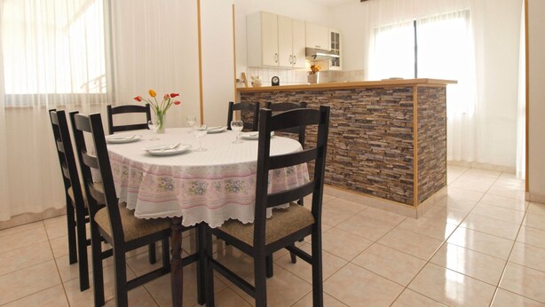 Een eethoek met een ronde tafel, zes stoelen en een vaas met bloemen. Er is ook een keuken met een betegelde achterwand en een aanrecht.