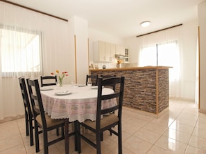 Uma sala de jantar com uma mesa redonda, seis cadeiras e um vaso com flores. Há também uma cozinha com painel de fundo de pedra e bancada.