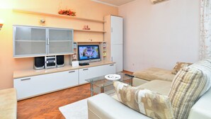 Um espaço de estar compacto com sofá, mesa de centro de vidro, TV e um armário branco com sistema de som embutido.