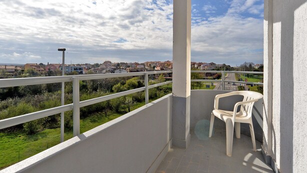 Uma varanda com guarda-corpo branco, uma cadeira de plástico e vista para um bairro residencial com casas e vegetação.