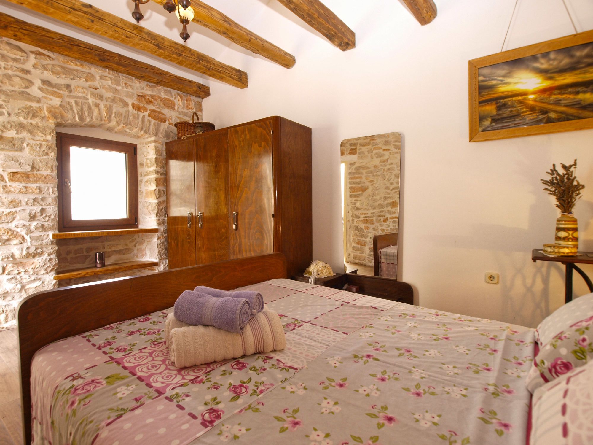 Ein Schlafzimmer mit Steinwand, Holzbalkendecke, einem großen Bett mit Blumenbettwäsche, einem Holzschrank und einem Gemälde einer Sonnenuntergangslandschaft.
