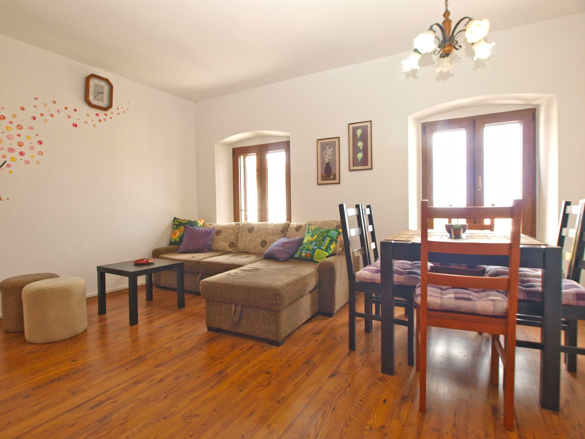 A living room with a sectional sofa, a dining table with chairs, a small ottoman, and a chandelier.