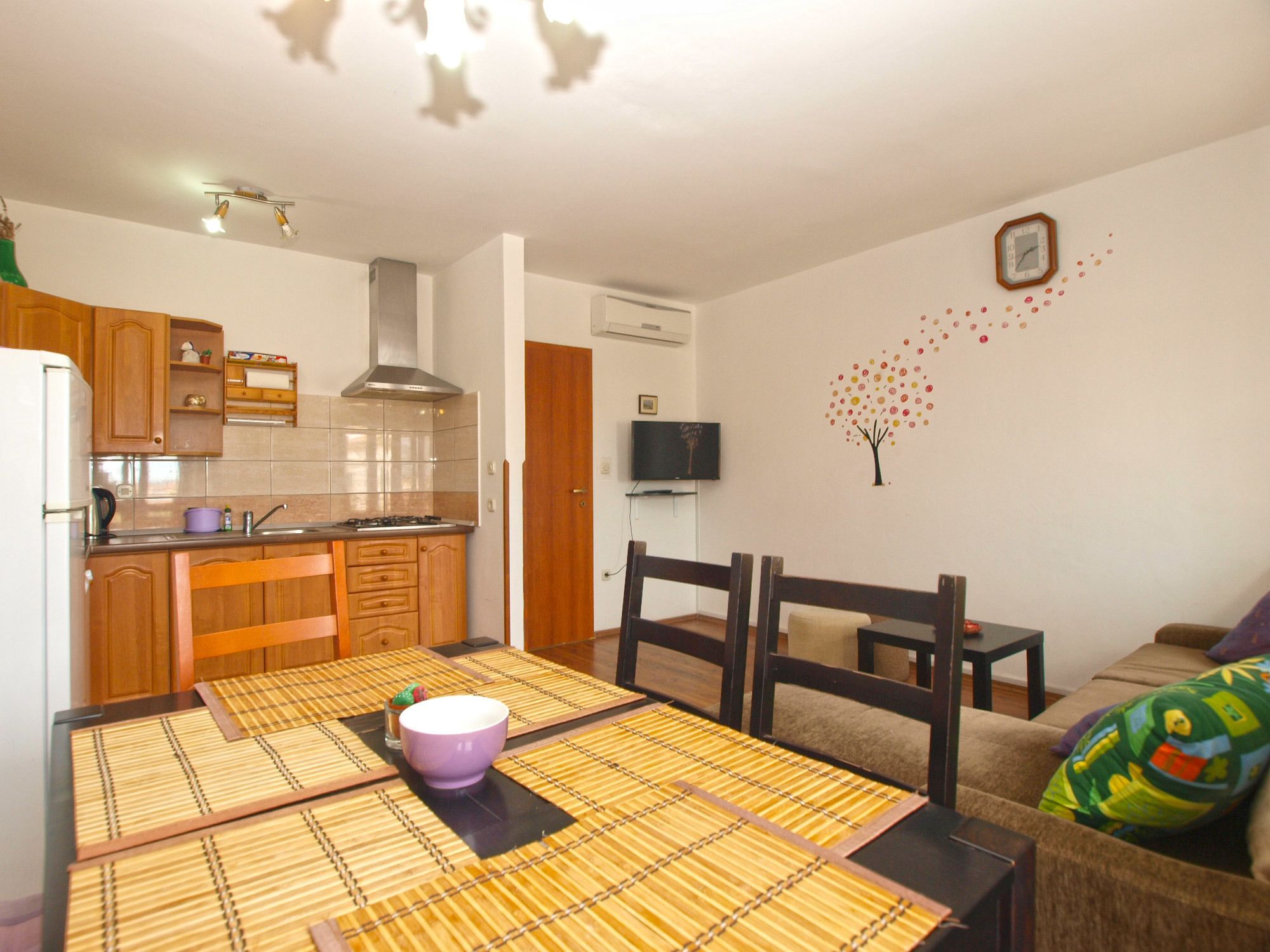 A compact kitchen with wooden cabinets, a gas stove, and a microwave. There's a dining table with a placemat and a purple bowl. A TV is mounted on the wall, and there's a small plant on the counter.