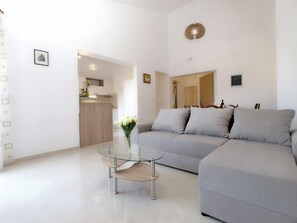 Una sala moderna con un sofá gris, una mesa de centro de cristal y un jarrón con flores. Se ve un área de cocina al fondo.