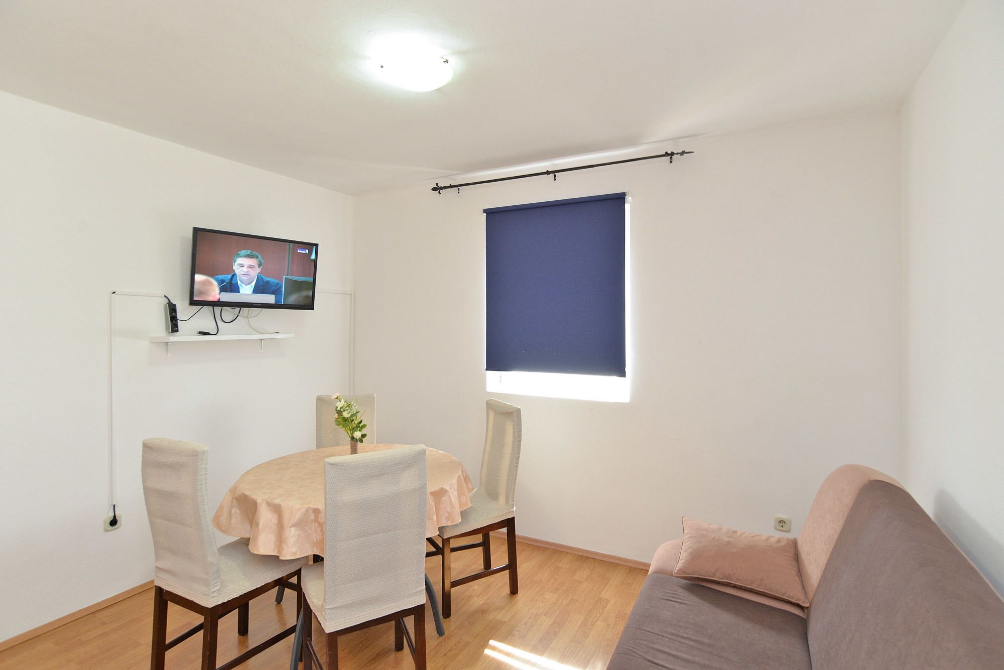 A living room with a television on the wall, a dining table with chairs, a sofa, and a window with a blind.