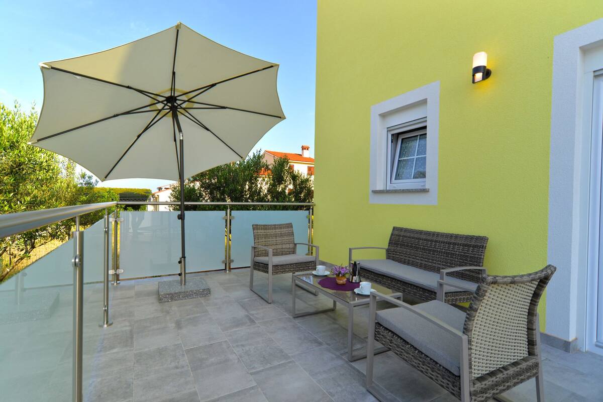 A balcony with a glass railing, a beige umbrella, wicker furniture, and a small table with cups and a vase.