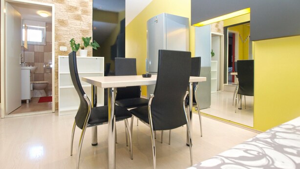 A modern dining area with a white table and black chairs, a kitchen in the background, and a yellow accent wall. - Apartment 1623 (Fazana)