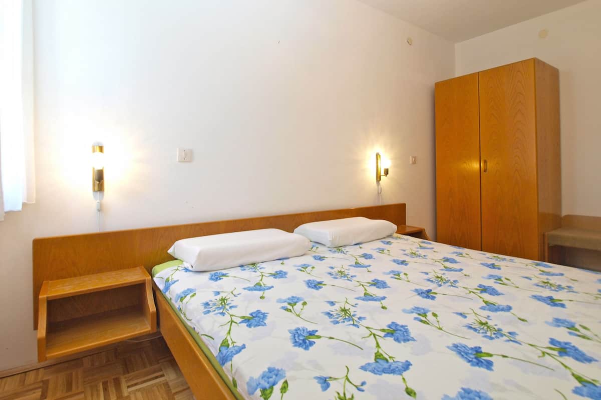 A bedroom with a wooden headboard, a bed with a floral bedspread, a nightstand, a wooden wardrobe, and two wall-mounted light fixtures.