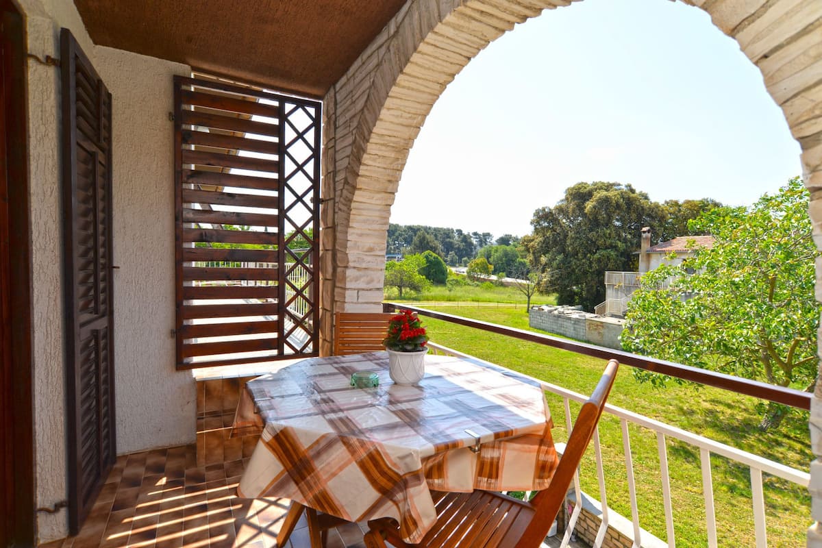 A balcony with a table and chairs, a view of greenery and buildings, and a stone archway.
