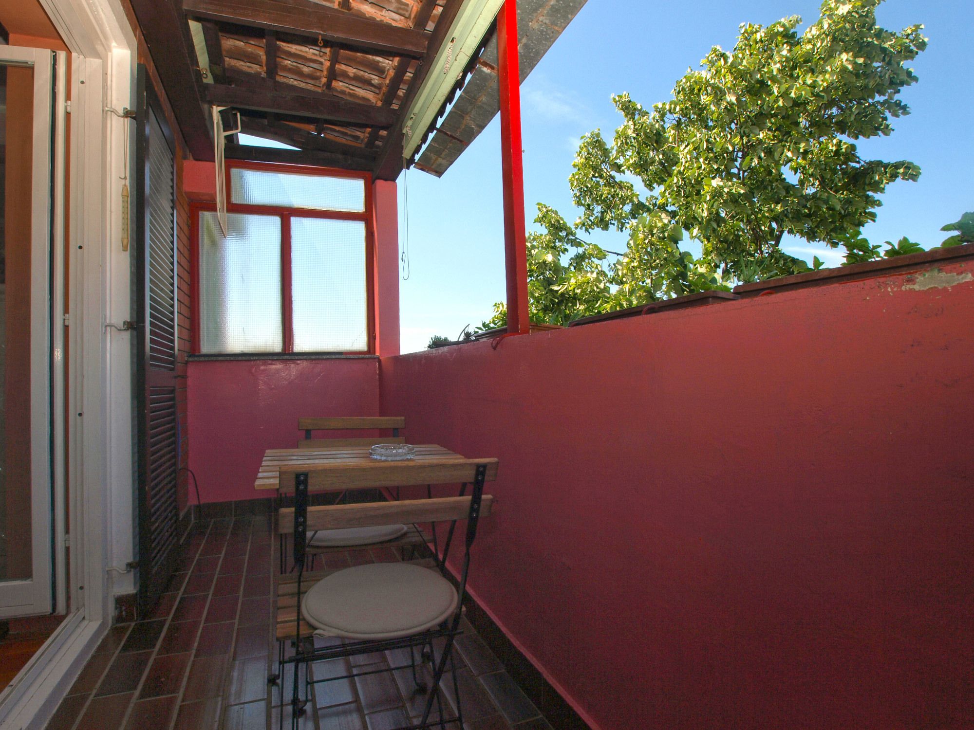 A small balcony with a wooden table and chairs, a window with blinds, and a red wall.