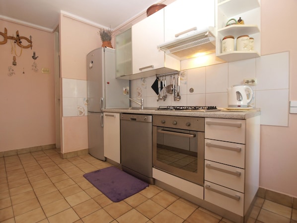 Compact kitchen with refrigerator, dishwasher, oven, and sink, featuring a tiled backsplash and countertop with a kettle and storage.
